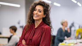 Junge Frau mit lockigem Haar und bordeauxroter Bluse sitzt lächelnd in einem modernen Büroraum – Symbolbild für das MBA-Studium in Projekt- und Prozessmanagement an der Hochschule Burgenland.