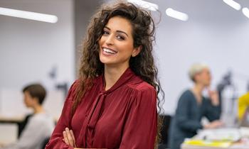 Junge Frau mit lockigem Haar und bordeauxroter Bluse sitzt lächelnd in einem modernen Büroraum – Symbolbild für das MBA-Studium in Projekt- und Prozessmanagement an der Hochschule Burgenland.