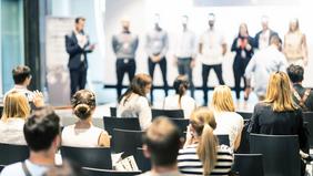 Studierende und Gäste sitzen in einem Veranstaltungsraum und folgen einer Präsentation auf der Bühne, bei der mehrere Personen stehen. Die Szene zeigt eine Preisverleihung oder eine Projektvorstellung – sinnbildlich für Innovation, Motivation und Sichtbarkeit im Rahmen der FH Burgenland Awards.