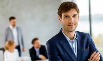 Junger Mann in dunkelblauem Sakko blickt freundlich in die Kamera, im Hintergrund unscharf mehrere Personen in einem modernen Besprechungsraum. Das Bild visualisiert Professionalität, Führungsanspruch und die Zielgruppe des MBA Unternehmensführung der FH Burgenland.