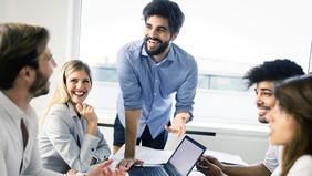 Fünf junge Menschen in einem modernen Büro in lebhafter Diskussion, lachend und mit Laptop – symbolisiert innovative Zusammenarbeit im Bereich Digitalisierung und Management.