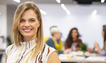 Diploma of Advanced Studies in Change Leadership und Coaching Lächelnde blonde Frau in einem hellen, gemusterten Oberteil steht in einem modernen Büro mit unscharfem Hintergrund, in dem mehrere Personen an einem Tisch arbeiten. Das Bild vermittelt Professionalität und Offenheit – passend zur Weiterbildung in Change Leadership und Coaching.