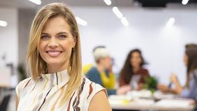 Lächelnde blonde Frau in einem hellen, gemusterten Oberteil steht in einem modernen Büro mit unscharfem Hintergrund, in dem mehrere Personen an einem Tisch arbeiten. Das Bild vermittelt Professionalität und Offenheit – passend zur Weiterbildung in Change Leadership und Coaching.