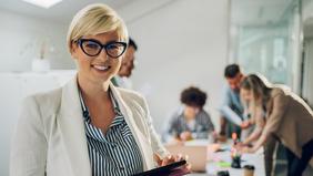 Junge Frau mit Tablet lächelt in die Kamera, im Hintergrund arbeiten mehrere Personen gemeinsam an einem Tisch – Symbolbild für das berufsbegleitende MBA-Studium IT-Management an der Hochschule Burgenland.
