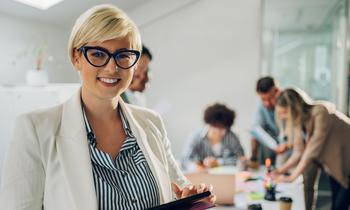 Junge Frau mit Tablet lächelt in die Kamera, im Hintergrund arbeiten mehrere Personen gemeinsam an einem Tisch – Symbolbild für das berufsbegleitende MBA-Studium IT-Management an der Hochschule Burgenland.