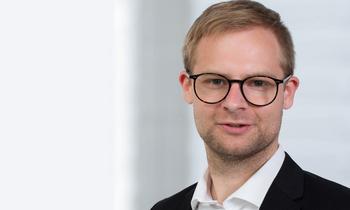 Porträt von Christopher Hillebrand, MBA-Student an der FH Burgenland, in einem hellen Setting. Er trägt eine schwarze Brille und einen dunklen Anzug mit weißem Hemd – sinnbildlich für Engagement, Innovationsgeist und die Verbindung von IT und Leadership.