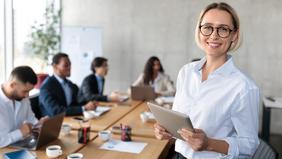 Diploma of Advanced Studies in Business Management und New Business Development Junge Frau mit Brille und Tablet in einem modernen Besprechungsraum, im Hintergrund ein dynamisches Team bei der Zusammenarbeit – sinnbildlich für Innovation und neue Geschäftsentwicklung im DAS Business Management und New Business Development der FH Burgenland.