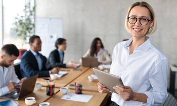 Junge Frau mit Brille und Tablet in einem modernen Besprechungsraum, im Hintergrund ein dynamisches Team bei der Zusammenarbeit – sinnbildlich für Innovation und neue Geschäftsentwicklung im DAS Business Management und New Business Development der FH Burgenland.