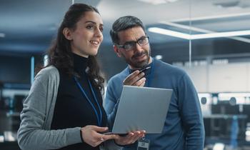 Zwei IT-Fachkräfte, eine Frau und ein Mann mit Tablet, stehen in moderner Arbeitsumgebung und diskutieren – Symbolbild für den MBA Informationssicherheit und IT-Risikomanagement an der Hochschule Burgenland.