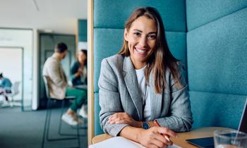 MA (CE) Personalmarketing und Employer Branding Junge Frau mit Notizbuch und Laptop sitzt lächelnd an einem Tisch in modernem Büro – steht für zeitgemäßes Arbeiten im Personalmarketing.