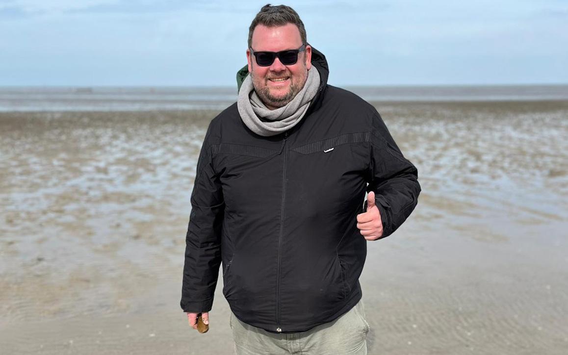 Marc Schachteli am Strand bei Wind, mit Sonnenbrille und Jacke, lächelt in die Kamera