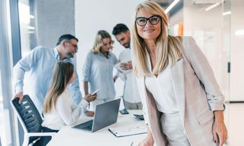 Selbstbewusste Frau in Businesskleidung blickt lächelnd in die Kamera, im Hintergrund ein Team im Gespräch – Sinnbild für moderne Führung und Teamarbeit im beruflichen Kontext.