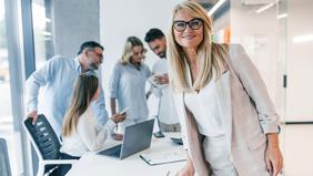 Selbstbewusste Frau in Businesskleidung blickt lächelnd in die Kamera, im Hintergrund ein Team im Gespräch – Sinnbild für moderne Führung und Teamarbeit im beruflichen Kontext.