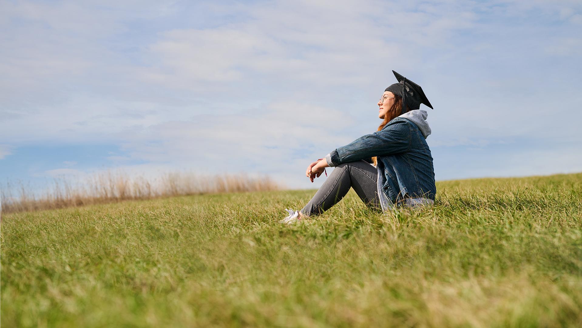 Studierende mit Absolventenhut sitzt entspannt auf einer Wiese und blickt in den Himmel – Symbol für individuelle Betreuung, persönliche Entwicklung und Zukunftsperspektiven an der Hochschule Burgenland.