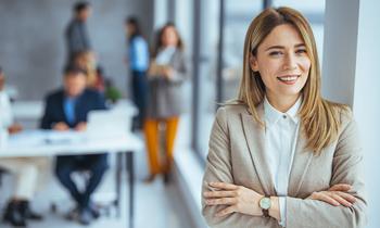 Junge Frau mit langen blonden Haaren und beigefarbenem Blazer steht selbstbewusst lächelnd in einem modernen Büro – Symbolbild für das MBA-Studium in Digital Business Management an der Hochschule Burgenland.