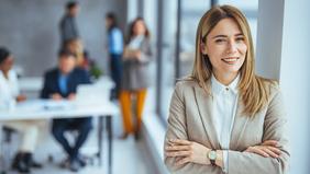 Junge Frau mit langen blonden Haaren und beigefarbenem Blazer steht selbstbewusst lächelnd in einem modernen Büro – Symbolbild für das MBA-Studium in Digital Business Management an der Hochschule Burgenland.
