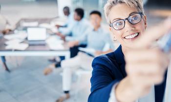 Lächelnde Führungskraft in Business-Outfit macht ein Selfie mit ihrem Team im Hintergrund – Symbol für moderne Leadership, Teamspirit und innovative Managementansätze.