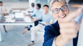 Lächelnde Führungskraft in Business-Outfit macht ein Selfie mit ihrem Team im Hintergrund – Symbol für moderne Leadership, Teamspirit und innovative Managementansätze.