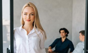 Junge Frau steht in einem modernen Büro und blickt direkt in die Kamera. Im Hintergrund sitzen zwei Männer im Gespräch. Das Bild symbolisiert berufsbegleitende Weiterbildung im Bereich Wirtschaftspsychologie – auch für Praktiker*innen ohne Matura.