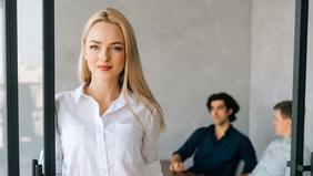MBA Wirtschaftspsychologie Junge Frau steht in einem modernen Büro und blickt direkt in die Kamera. Im Hintergrund sitzen zwei Männer im Gespräch. Das Bild symbolisiert berufsbegleitende Weiterbildung im Bereich Wirtschaftspsychologie – auch für Praktiker*innen ohne Matura.