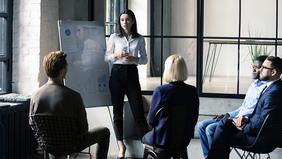 Junge Frau steht vor Flipchart und spricht zu einer Gruppe von Menschen – veranschaulicht den MBA-Lehrgang Wirtschaftsmediation an der FH Burgenland mit Fokus auf Konfliktlösung und Beratungsprozesse.