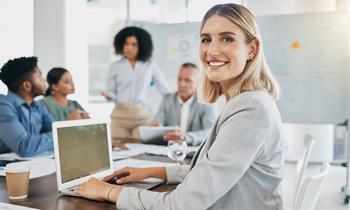 Junge Frau im Business-Outfit sitzt an einem Konferenztisch mit Laptop und lächelt in die Kamera, während im Hintergrund ein kreatives Team an Markenstrategien arbeitet – stellvertretend für den MBA Brand Management der FH Burgenland.