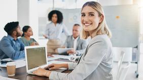 MBA Brand Management Junge Frau im Business-Outfit sitzt an einem Konferenztisch mit Laptop und lächelt in die Kamera, während im Hintergrund ein kreatives Team an Markenstrategien arbeitet – stellvertretend für den MBA Brand Management der FH Burgenland.