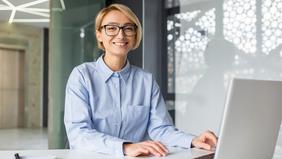 Junge Frau mit Brille sitzt freundlich lächelnd an einem Schreibtisch mit Laptop – Symbolbild für das berufsbegleitende MBA-Programm in Compliance, ESG und Risikomanagement an der Hochschule Burgenland.