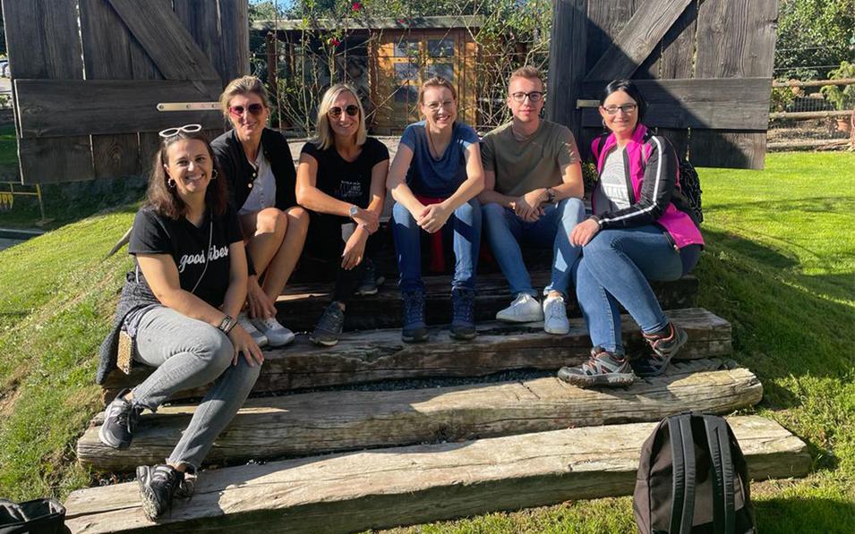 Mitarbeitende der FH Burgenland Weiterbildung sitzen entspannt auf Holzstufen vor einem dekorativen Tor im Grünen – ein Moment des gemeinsamen Erlebens.