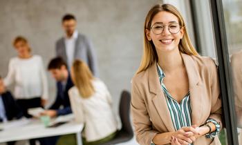Junge Frau mit Brille und Blazer lächelt selbstbewusst in die Kamera. Im Hintergrund diskutiert eine Gruppe von Personen an einem Tisch – Symbolbild für modernes, praxisorientiertes Fernstudium im agilen Management.
