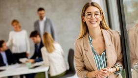 Junge Frau mit Brille und Blazer lächelt selbstbewusst in die Kamera. Im Hintergrund diskutiert eine Gruppe von Personen an einem Tisch – Symbolbild für modernes, praxisorientiertes Fernstudium im agilen Management.