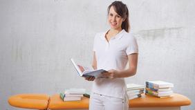 MSc (CE) Advanced Physiotherapy & Management Junge Frau in weißer Kleidung steht mit einem geöffneten Buch in der Hand vor einer Liege und mehreren Bücherstapeln – symbolisch für den MSc (CE) Advanced Physiotherapy & Management der FH Burgenland.
