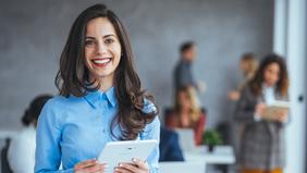 Junge Frau im Business-Outfit hält ein Tablet und lächelt in die Kamera. Im Hintergrund stehen mehrere Personen in einem modernen Büroraum – Symbolbild für digitales Fernstudium im Bereich E-Commerce und Online-Marketing.