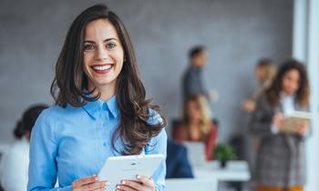 Junge Frau im Business-Outfit hält ein Tablet und lächelt in die Kamera. Im Hintergrund stehen mehrere Personen in einem modernen Büroraum – Symbolbild für digitales Fernstudium im Bereich E-Commerce und Online-Marketing.