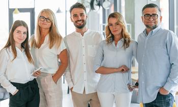 Fünf Erwachsene unterschiedlichen Alters stehen nebeneinander in heller, moderner Büroumgebung. Sie blicken selbstbewusst in die Kamera – sinnbildlich für die Zielgruppe der neuen Masterprogramme an der Hochschule Burgenland, die sich nun auch ohne Matura oder akademischen Abschluss weiterbilden kann.