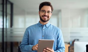 Junger Mann steht in einem modernen Büro und hält ein Tablet in der Hand. Er lächelt freundlich in die Kamera. Das Bild vermittelt digitale Kompetenz und Aufgeschlossenheit – passend zum MBA Management und Wirtschaftsinformatik der FH Burgenland.