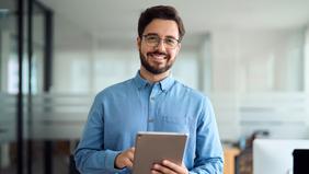 MBA Management und Wirtschaftsinformatik Junger Mann steht in einem modernen Büro und hält ein Tablet in der Hand. Er lächelt freundlich in die Kamera. Das Bild vermittelt digitale Kompetenz und Aufgeschlossenheit – passend zum MBA Management und Wirtschaftsinformatik der FH Burgenland.