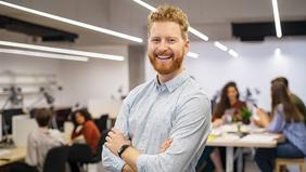 Junger Mann mit rotem Bart in hellem Hemd steht selbstbewusst lächelnd in einem modernen Büro mit offenem Arbeitsplatzkonzept. Im Hintergrund arbeiten mehrere Personen an Computern oder in Gruppen. Das Bild vermittelt Offenheit, Dynamik und Professionalität – passend zum MBA Business Management der FH Burgenland.