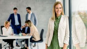 Junge Frau in beiger Jacke steht lächelnd an einer Glaswand, im Hintergrund arbeiten Personen in einem modernen Büro – symbolisiert nachhaltige Führungskompetenz im MBA Nachhaltigkeitsmanagement der FH Burgenland.