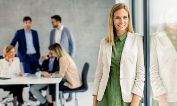 Junge Frau in beiger Jacke steht lächelnd an einer Glaswand, im Hintergrund arbeiten Personen in einem modernen Büro – symbolisiert nachhaltige Führungskompetenz im MBA Nachhaltigkeitsmanagement der FH Burgenland.