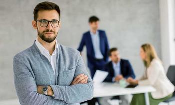Selbstbewusster junger Mann mit Brille steht mit verschränkten Armen im Vordergrund eines modernen Besprechungsraums, im Hintergrund diskutiert ein Team – Symbolbild für Führung, Risikobewertung und Krisenmanagement.