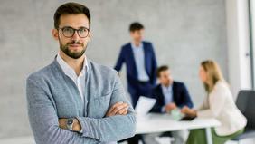 Selbstbewusster junger Mann mit Brille steht mit verschränkten Armen im Vordergrund eines modernen Besprechungsraums, im Hintergrund diskutiert ein Team – Symbolbild für Führung, Risikobewertung und Krisenmanagement.