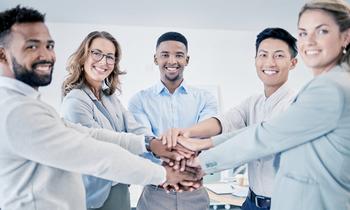 Fünf berufstätige Personen aus verschiedenen Hintergründen stehen im Halbkreis und legen lächelnd die Hände übereinander – Symbolbild für Teamarbeit und moderne Personalführung im MBA-Programm der FH Burgenland.