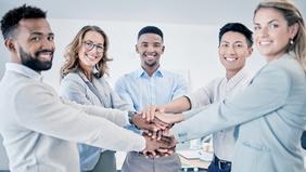 Fünf berufstätige Personen aus verschiedenen Hintergründen stehen im Halbkreis und legen lächelnd die Hände übereinander – Symbolbild für Teamarbeit und moderne Personalführung im MBA-Programm der FH Burgenland.