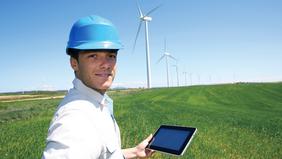 Mann mit Schutzhelm hält Tablet auf einem Feld vor Windrädern – Symbolbild für intelligente Energiesysteme und erneuerbare Energietechnologien.