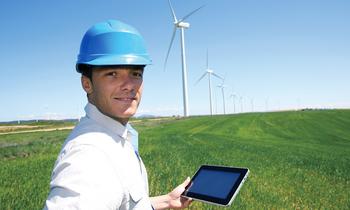 Mann mit Schutzhelm hält Tablet auf einem Feld vor Windrädern – Symbolbild für intelligente Energiesysteme und erneuerbare Energietechnologien.