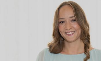 Anna Panzenböck lächelt freundlich in die Kamera. Sie trägt ein helles Oberteil und hat schulterlanges, gewelltes Haar. Der neutrale Hintergrund unterstreicht ihren professionellen Auftritt.