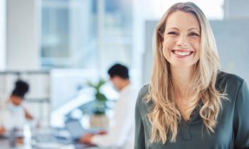Lächelnde Frau mit langen blonden Haaren in dunkler Bluse steht in einem modernen, hellen Büro mit Glasfront. Im Hintergrund arbeiten mehrere Personen an Laptops. Das Bild unterstreicht Offenheit, Aufbruch und neue Chancen – ideal zur Visualisierung der gesetzlichen Novelle zur akademischen Weiterbildung ab Juli 2024.
