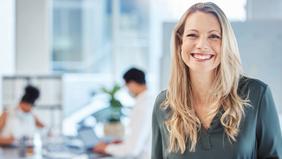 Lächelnde Frau mit langen blonden Haaren in dunkler Bluse steht in einem modernen, hellen Büro mit Glasfront. Im Hintergrund arbeiten mehrere Personen an Laptops. Das Bild unterstreicht Offenheit, Aufbruch und neue Chancen – ideal zur Visualisierung der gesetzlichen Novelle zur akademischen Weiterbildung ab Juli 2024.