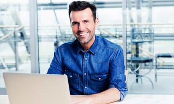 Lächelnder Mann in Freizeitbusiness-Kleidung sitzt vor einem Laptop in moderner Umgebung – symbolisiert den digitalen, ortsunabhängigen Zugang zum MBA IT-Management der FH Burgenland.